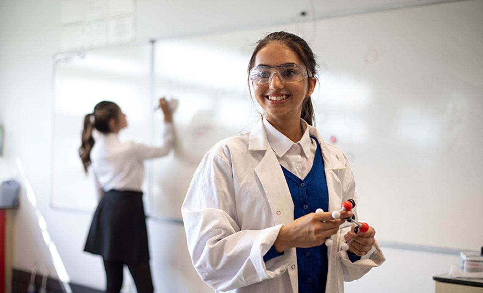 Kamala's pathway A teenage girl in a lab coat and protective glasses smiles to someone off camera. She is holding a DNA model in her hands.