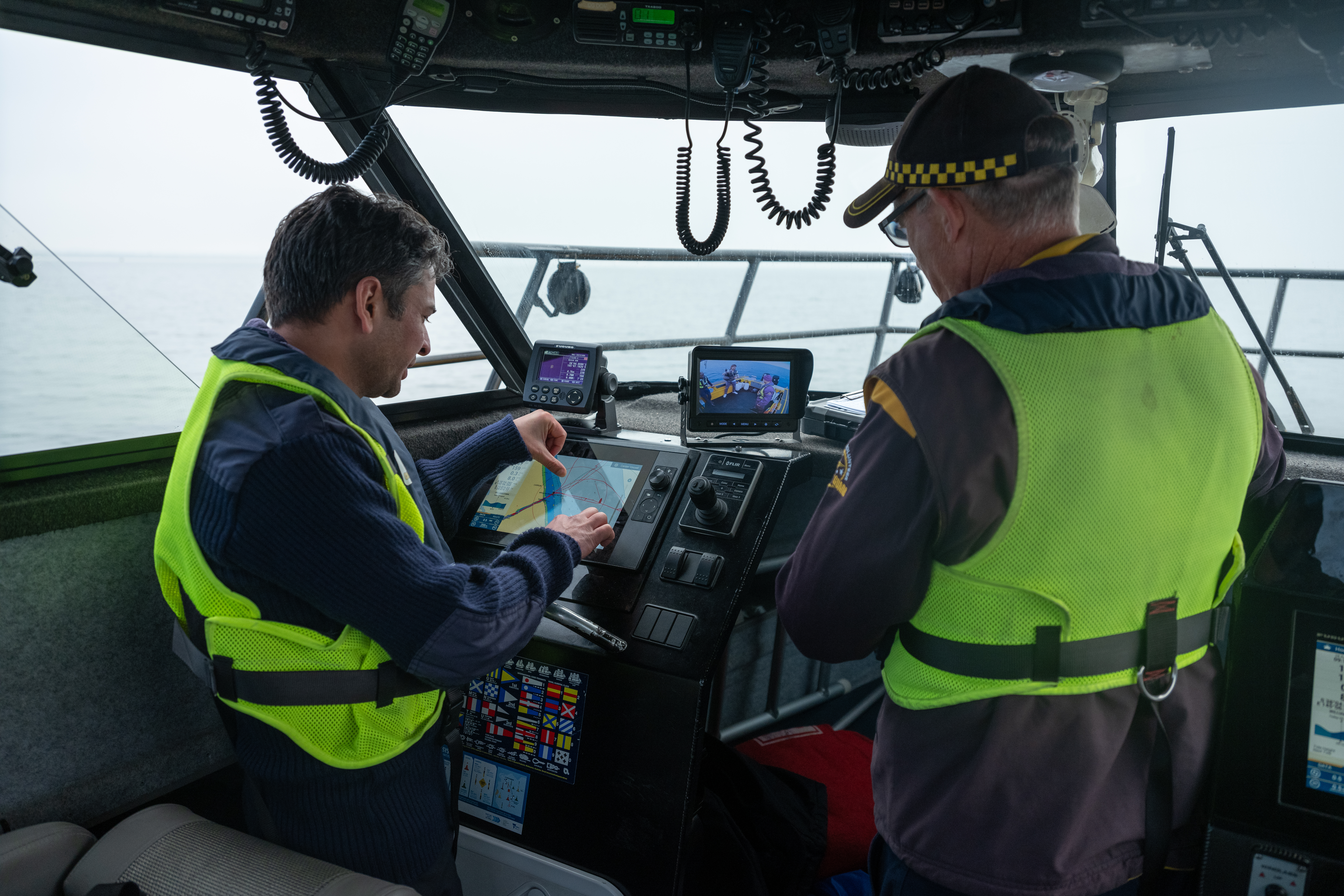 The image shows two men in life jackets, on a boat and using navigation equipment.