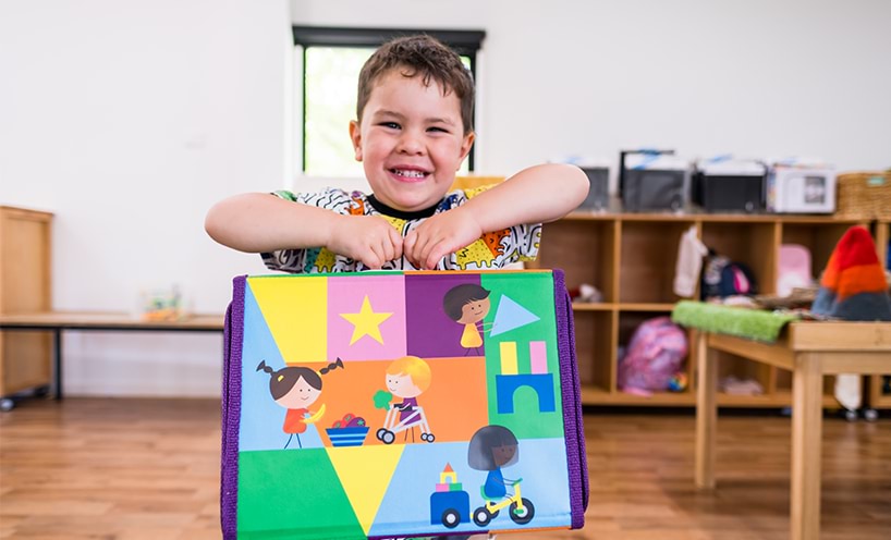 A kinder student is smiling at the camera as he holds his Kinder Kit up. The bag is covered in colourful illustrated, designs of children playing with toys.