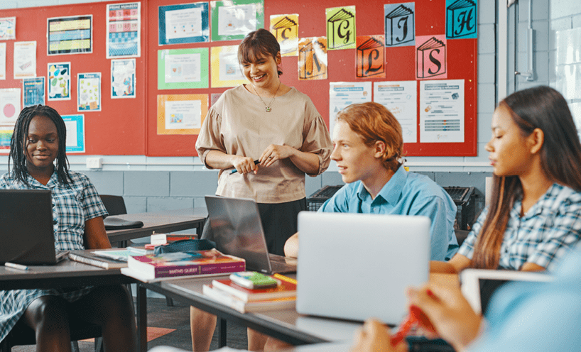 Female teacher standing in a classroom with three high school aged students on laptops