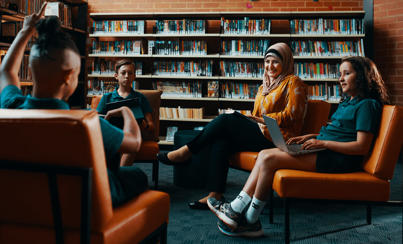 Female teacher with three primary school aged students sitting on chairs in a library