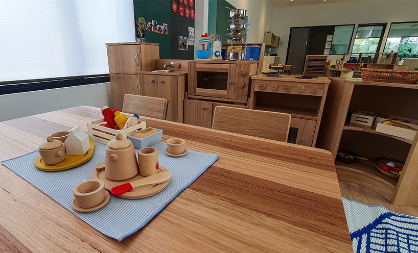 A wide-angled shot of a wooden table and kitchen. On the table sits wooden mugs, teapots and plates.