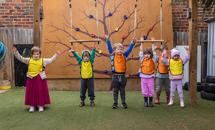 Six kinder students stand in a line with their arms in the air. They're outside beneath a swing set and in front of a small rock-climbing wall.