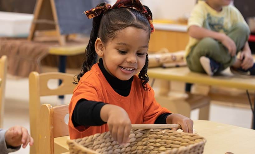 A kinder student in an orange shirt sits at a table. She is reaching into a basket and has a smile on her face.