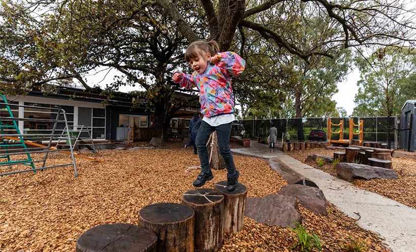 A preschool girl is playing outside on he playground. She is balancing on wooden stumps and she wears a colourful jumper.