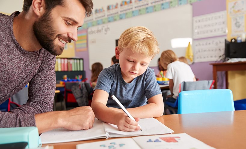 A male kindergarten teacher sits with a kinder student at a desk. The kinder student is writing in a booklet.