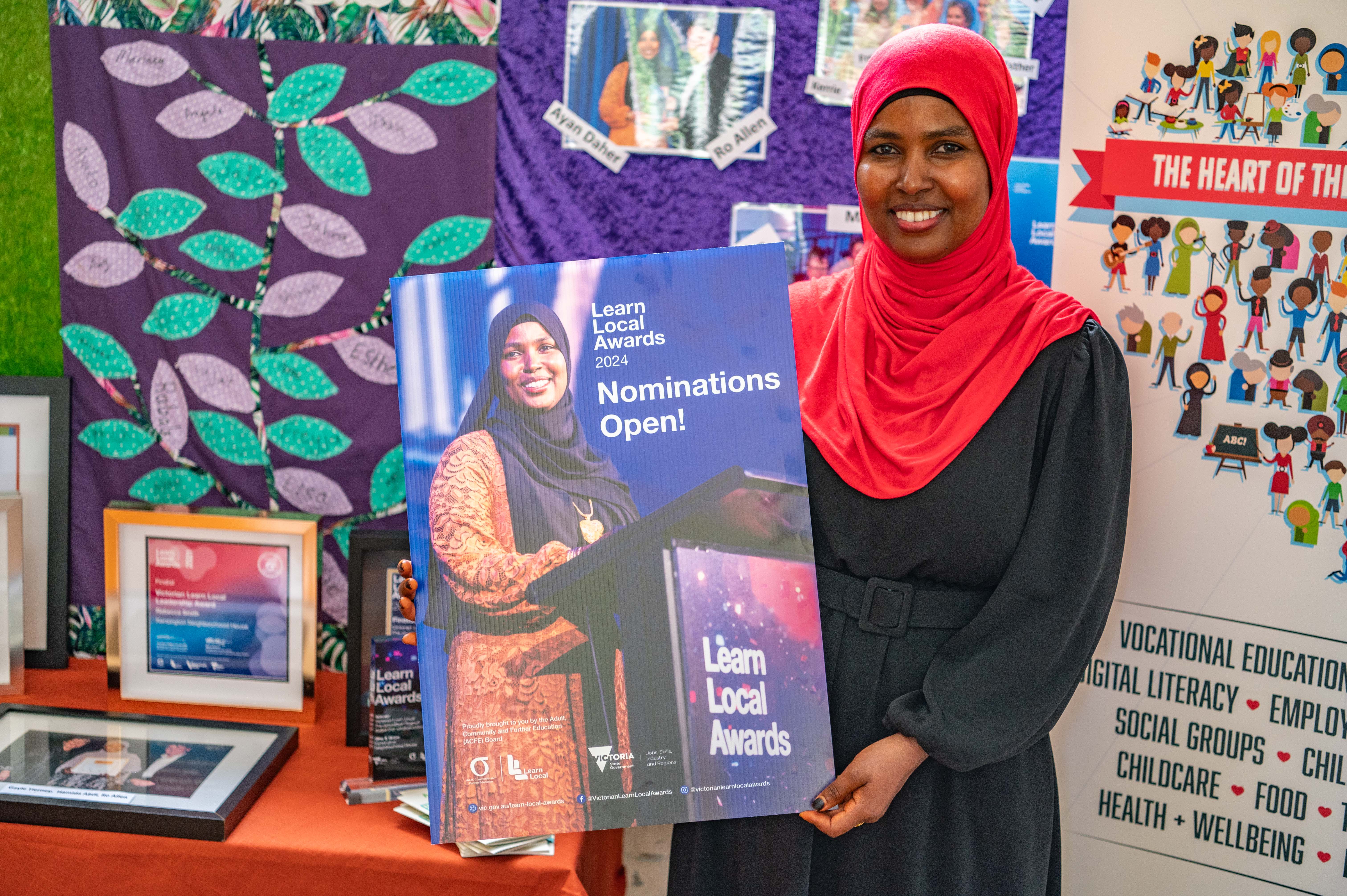 2023 Learn local award winner Ayan Daher holding up a poster of the Learn Local Awards programs for nominations open for 2024 standing in front of event stall promotion wall