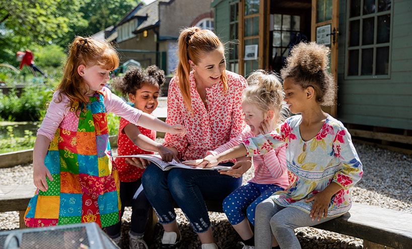 A kinder teacher sits with a group of children outside. She is reading a picture book to the children and they are all smiling.