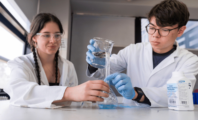 A male and female student in lab coats and glasses pour liquid into a beaker.