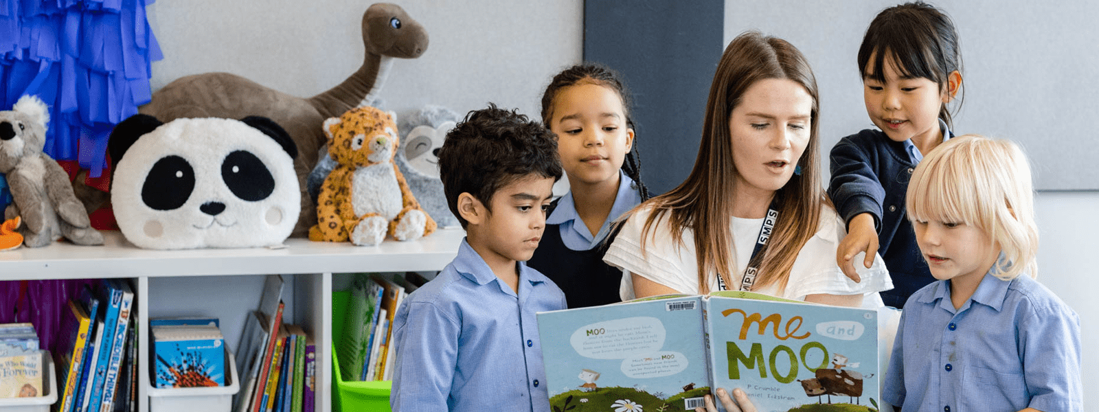 A teacher reads from a children's book with children gathered around her and surrounded by books and soft toys.