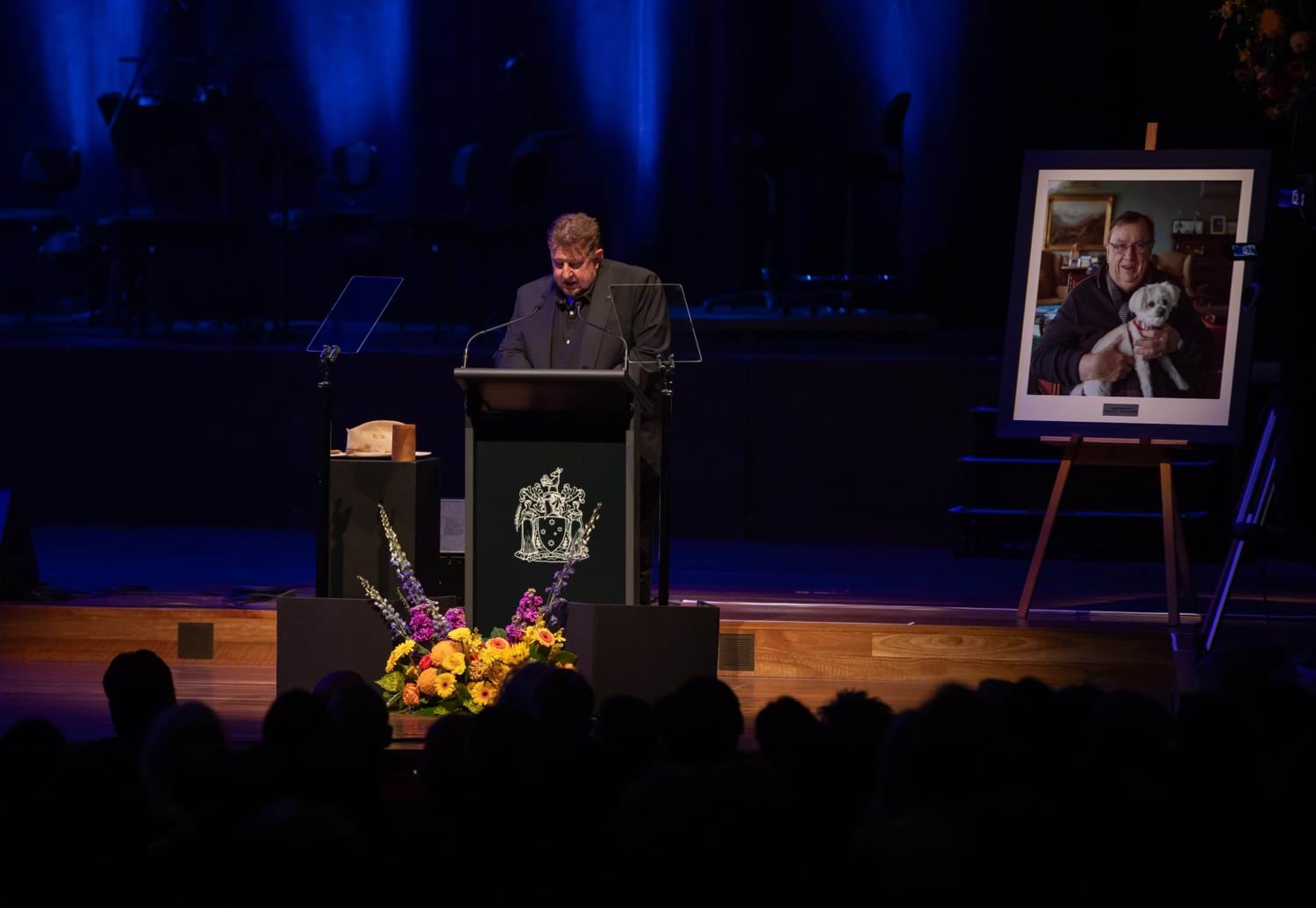 A man wearing black stands at a lectern on a stage with flowers and a framed photo of a man with a dog 