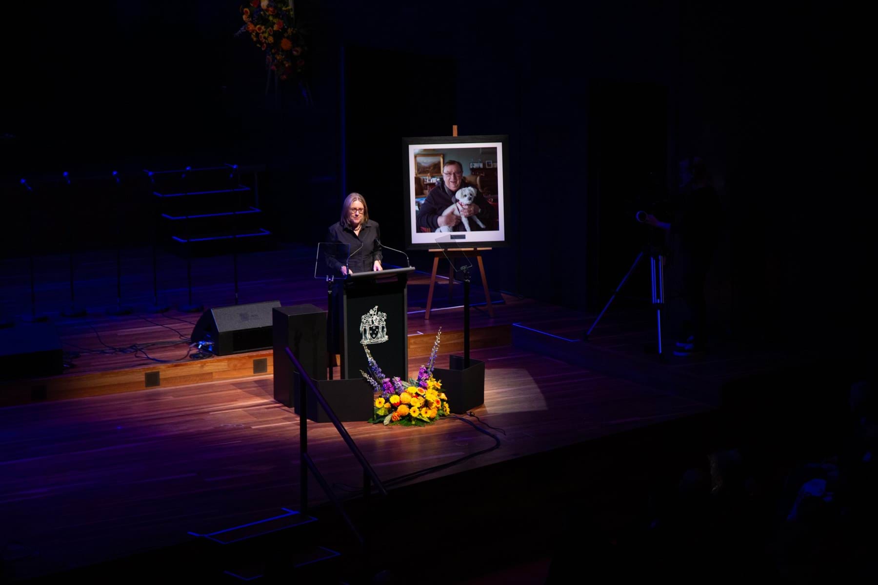 A women dressed in black stands at a podium on a stage with flowers