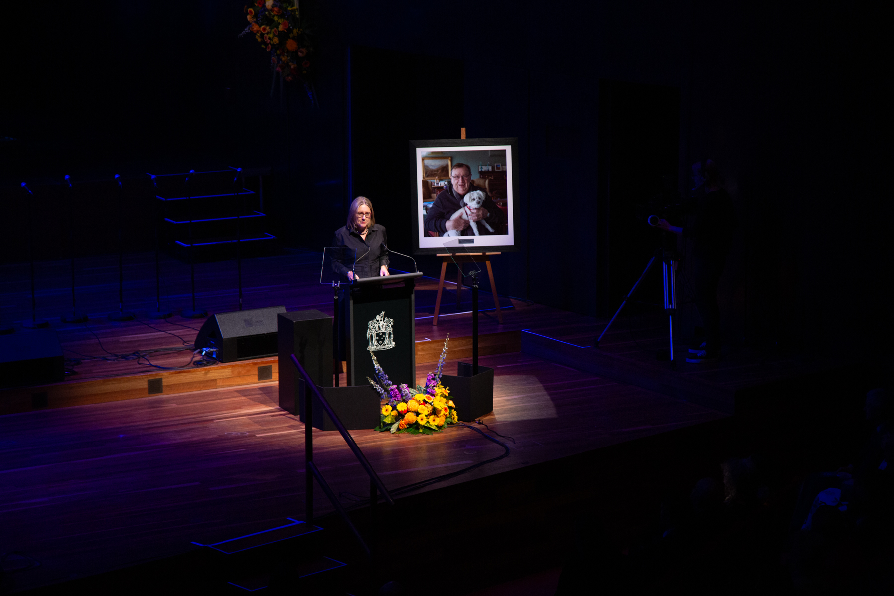 A women dressed in black stands at a podium on a stage with flowers