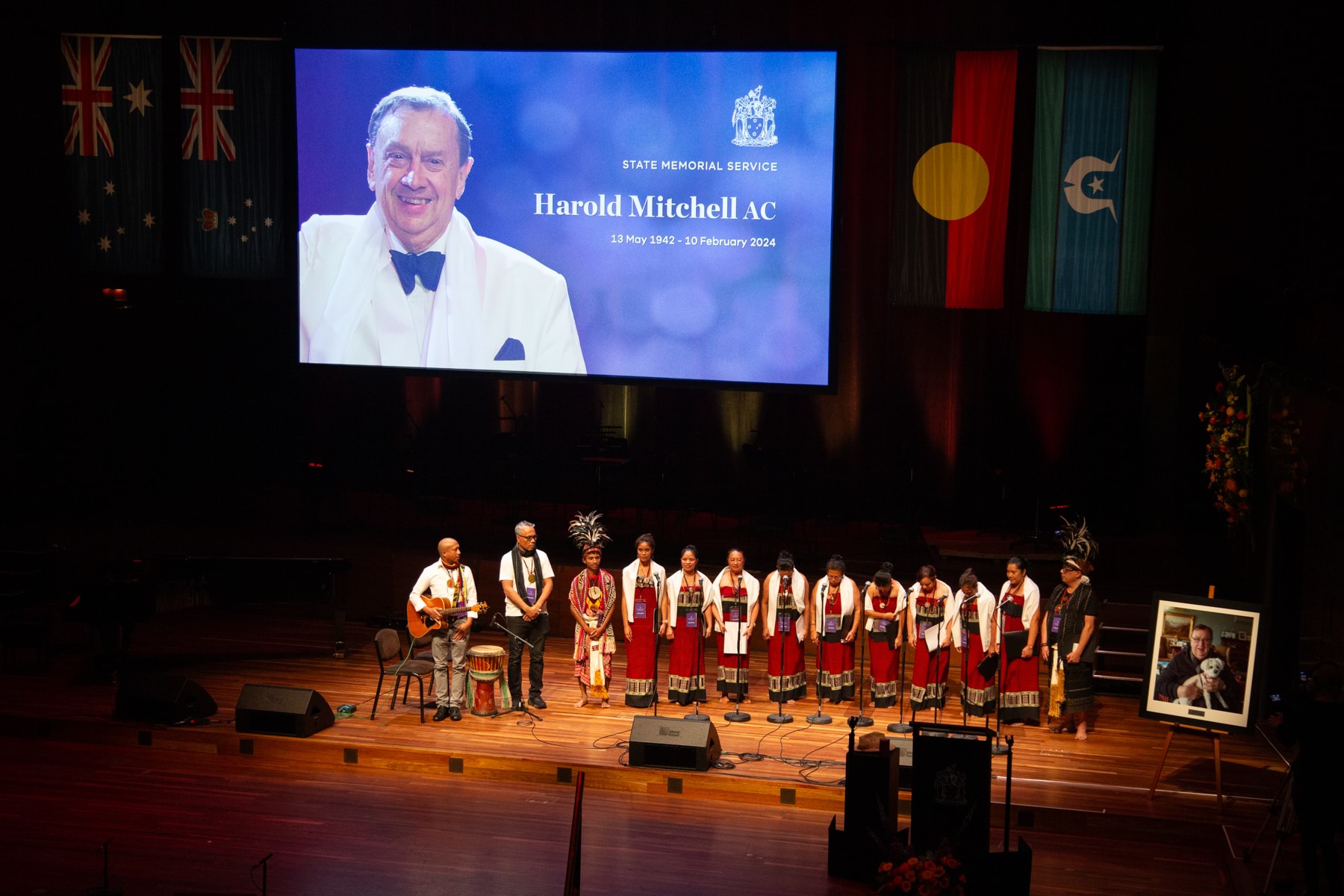 A group of musicians and singers wearing traditional dress on stage under a projection of a photo of a man with the words 'Harold Mitchell AC'