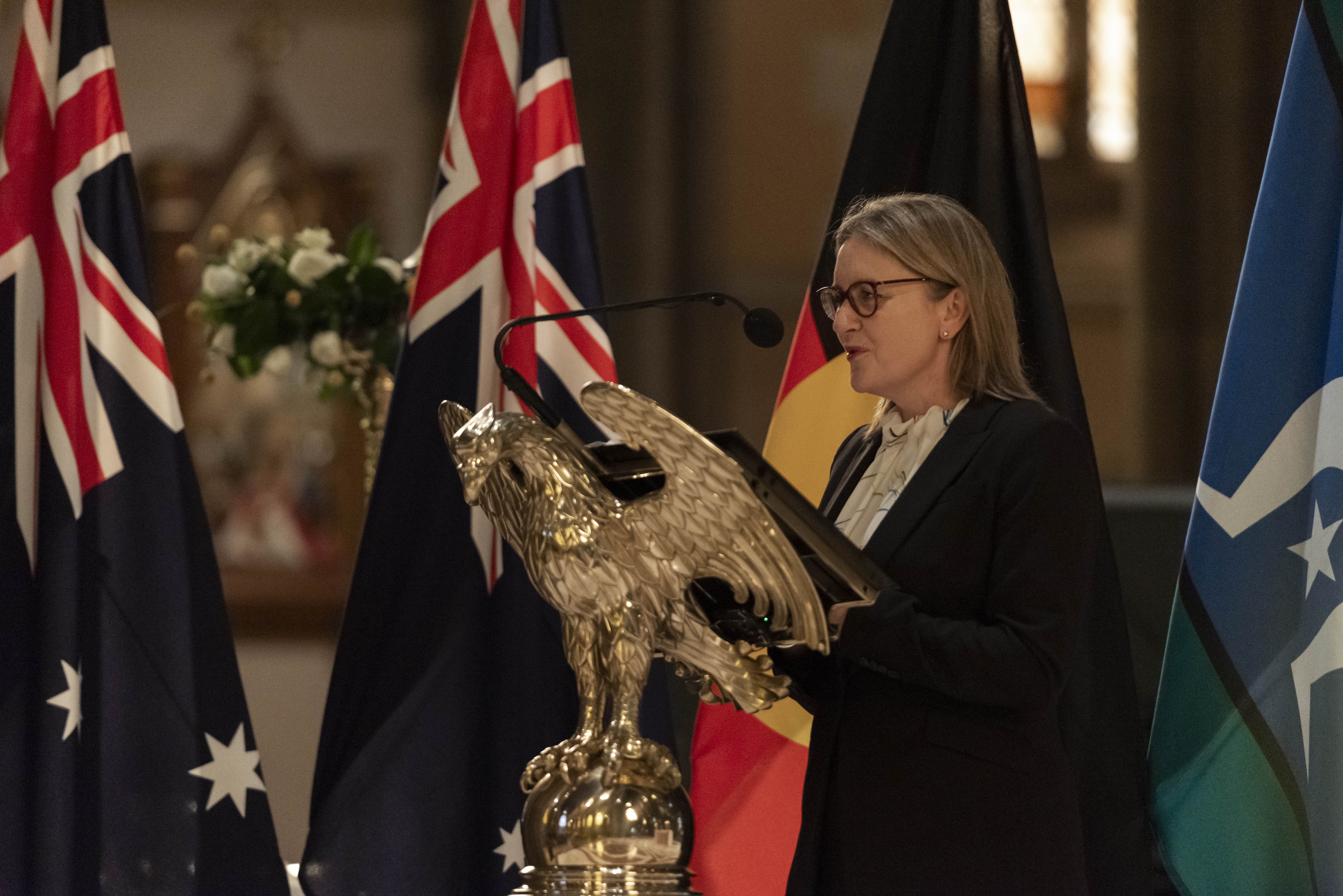 A woman with dark blonde hair in a white shirt and black blazer speaks into a microphone at a church pulpit, with the Australian, Victorian, Aboriginal and Torres Strait Islander flags in the background. 