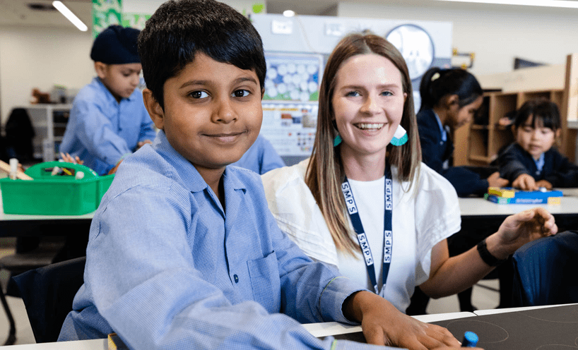 A male primary school student in a light blue shirt and a female teacher in a white shirt both smile at the camera.