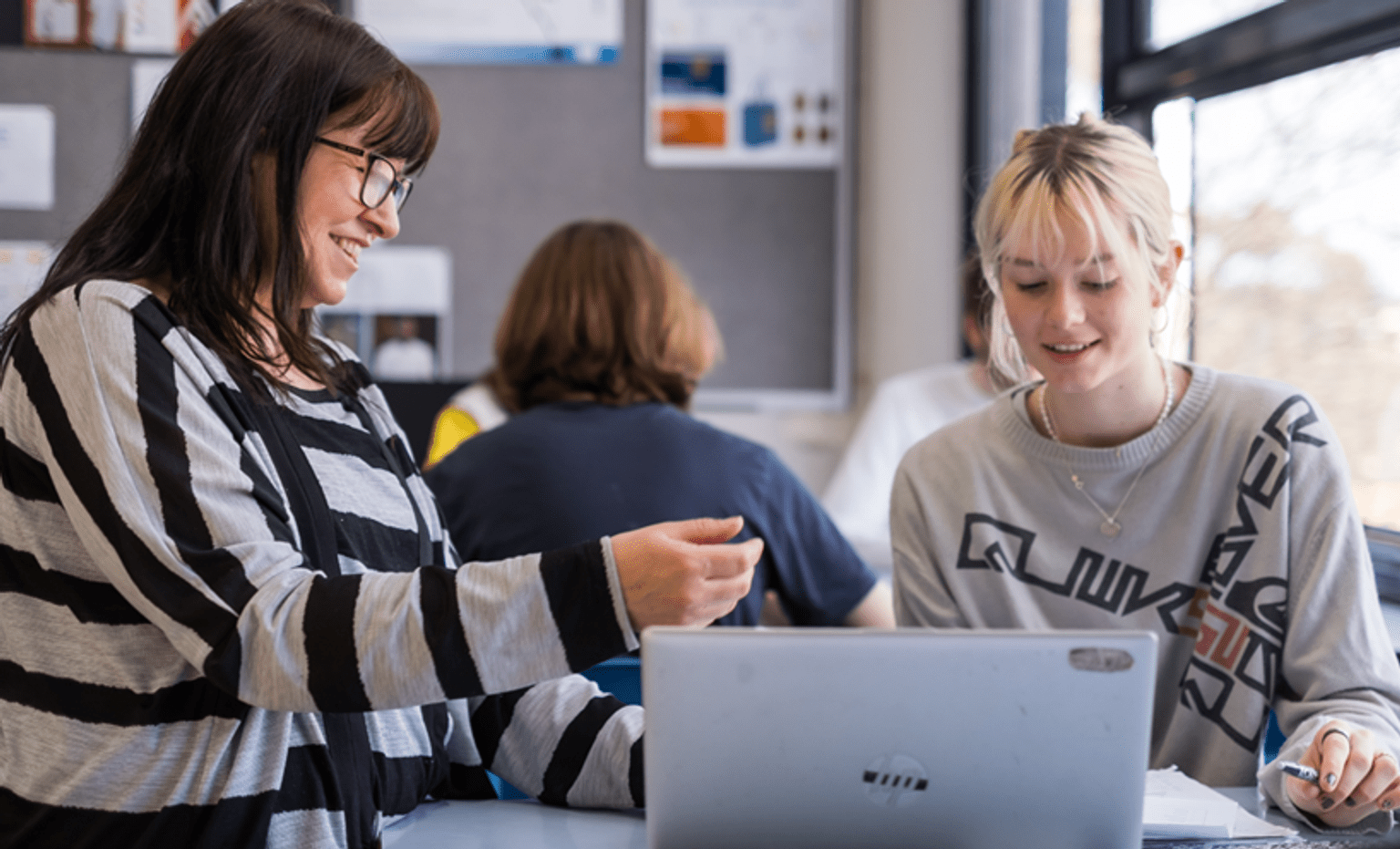 New teaching scholarship and other workforce supports  A female teacher in a grey and black striped shirt sits with a female high school student in a grey shirt. They are smiling down at a computer screen in front of them.