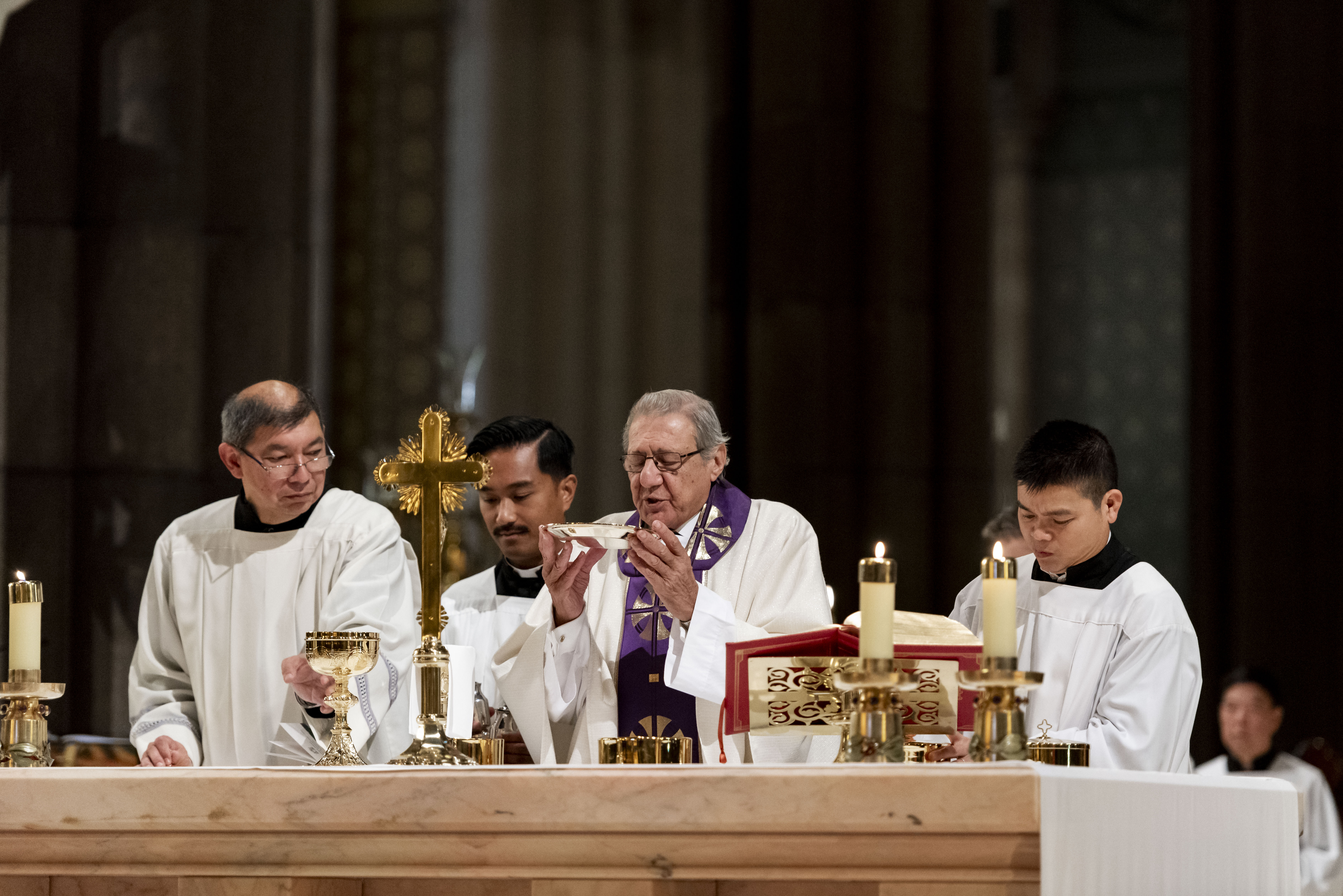 Four men wear white biblical dress and stand in front of an alter with a man positioned in the middle holding a silver communion plate.   