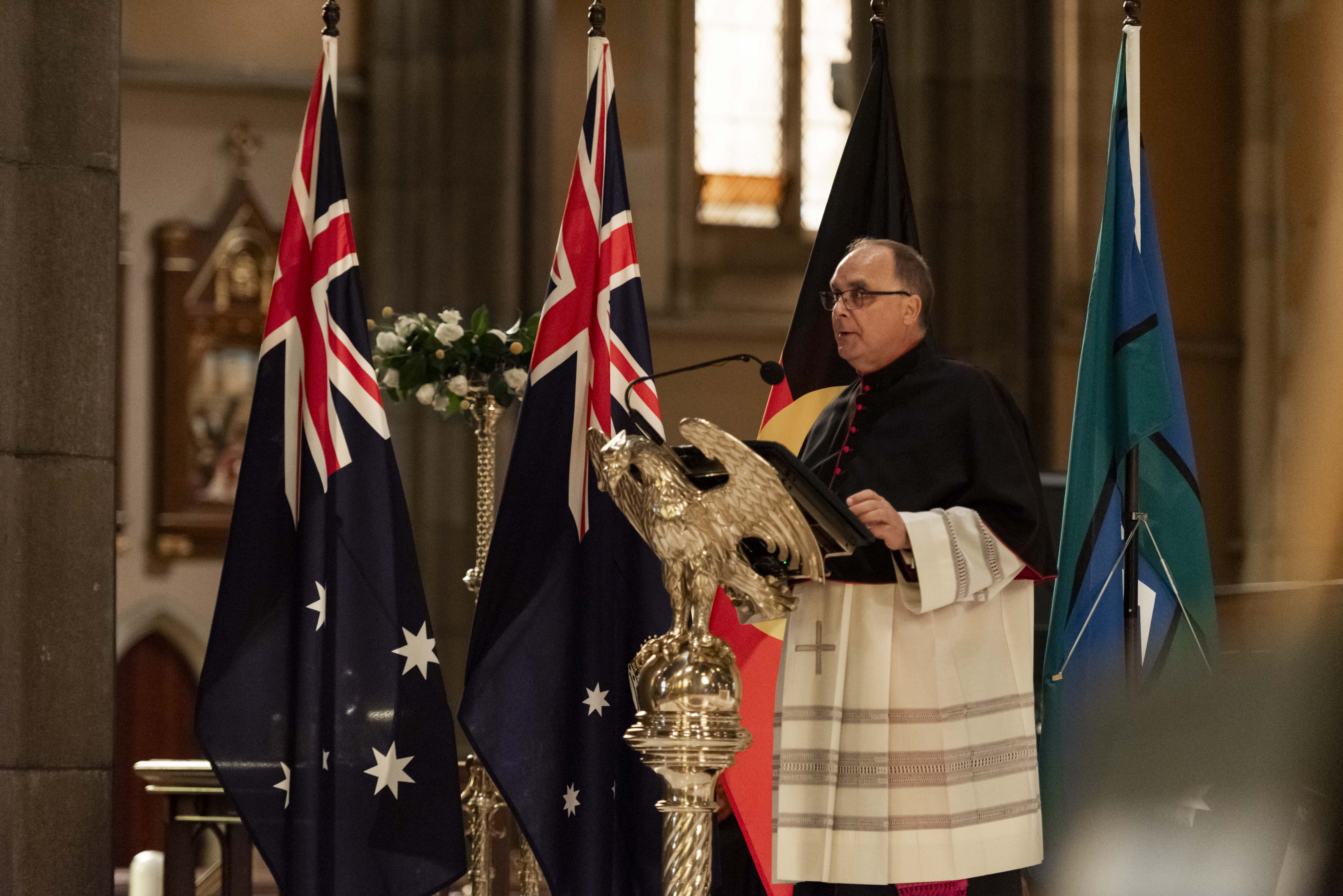 A man dressed in biblical garments addresses an audience from a church pulpit, with the Australian, Victorian, Aboriginal and Torres Strait Islander flags in the background. 