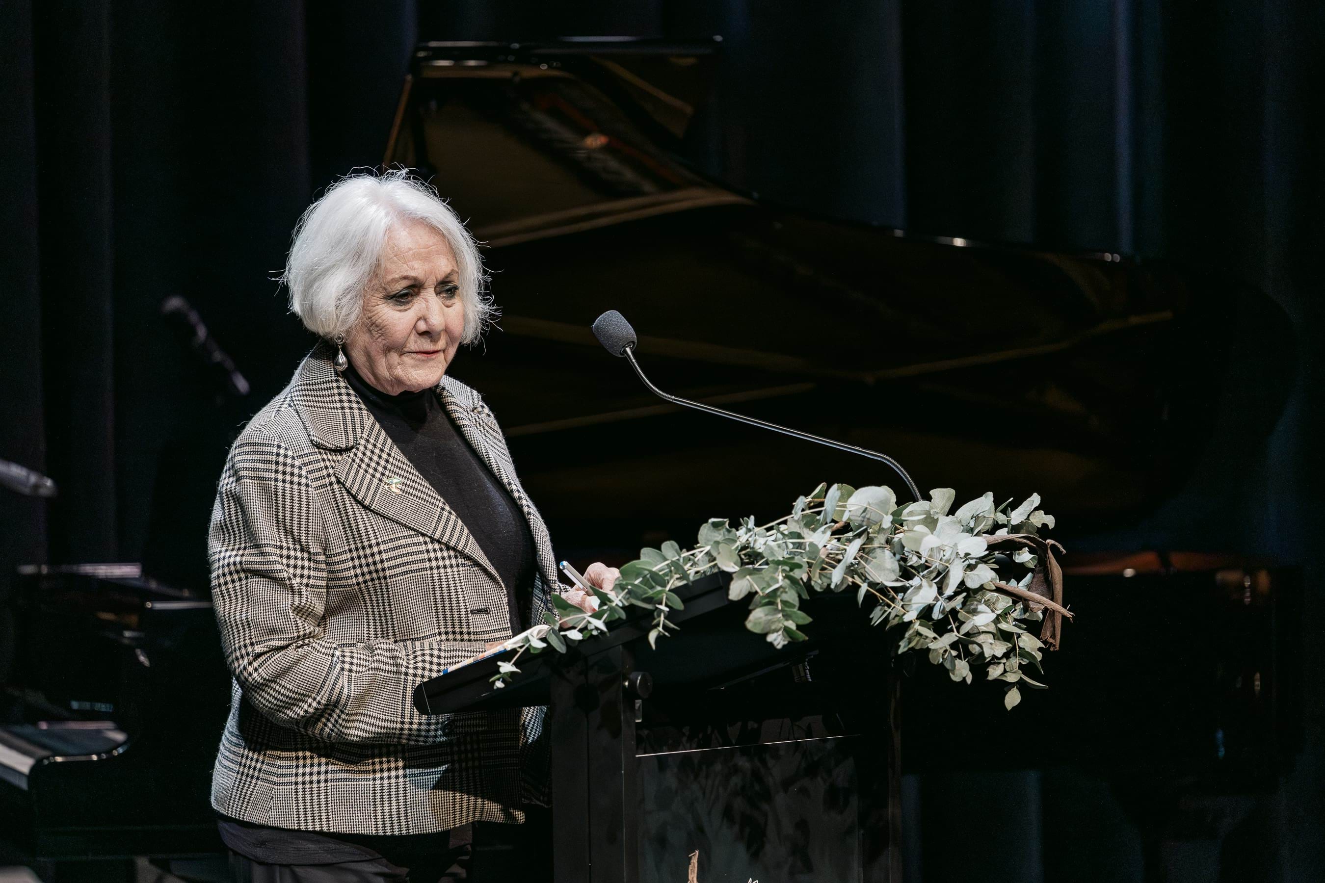 A lady with white hair and a grey jacket speaks at a podium with native flowers. 