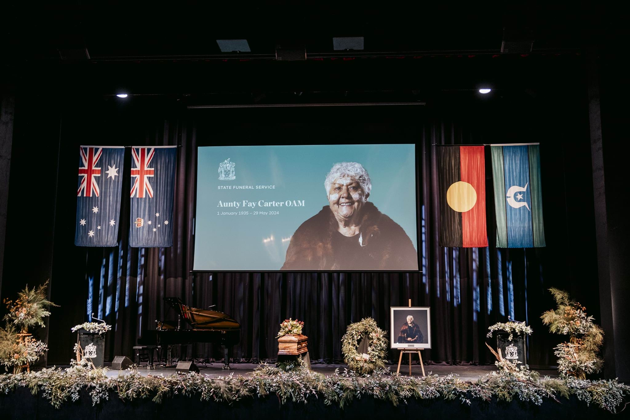 A stage with flowers, a coffin, four flags and a projection of an Aboriginal lady with the words 'Aunty Fay Carter OAM'