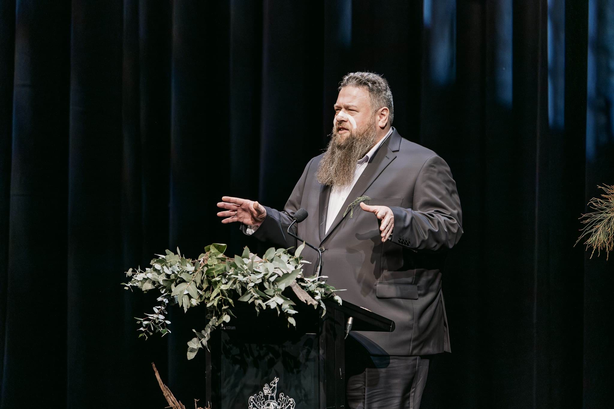 A man with a beard in a suit speaks at a podium with flowers. He has white face paint across his nose.