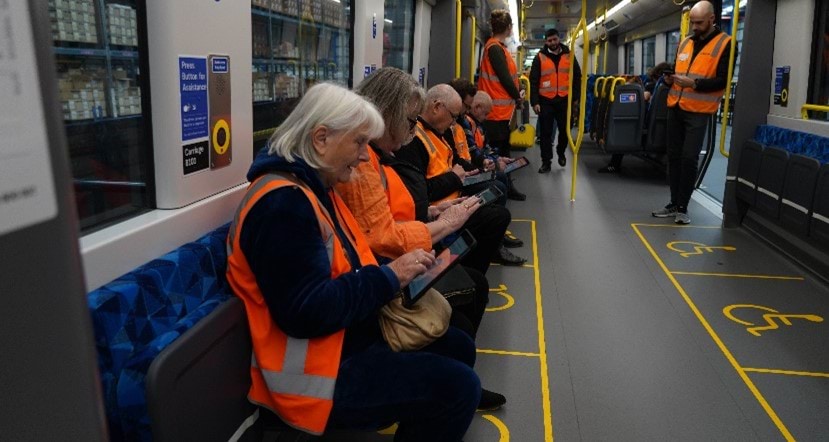 Photo of a group of people in orange vests sitting on a train mock-up and recording their feedback.