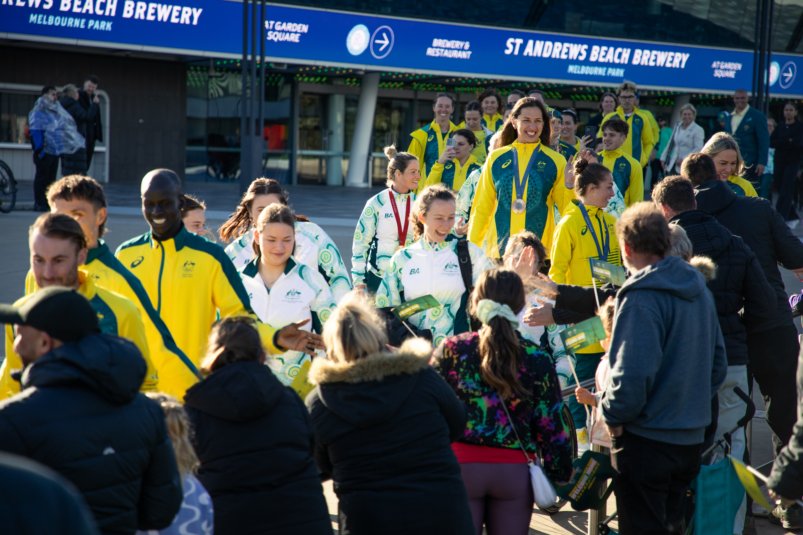 A line of athletes wearing matching tracksuits are smiling and waving as they walk down a walkway that is lined by people watching them. 