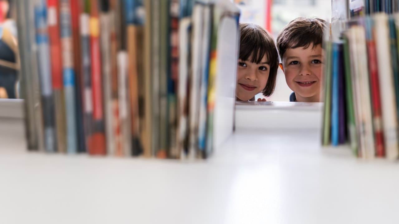 Two children smiling and peering through a bookcase.