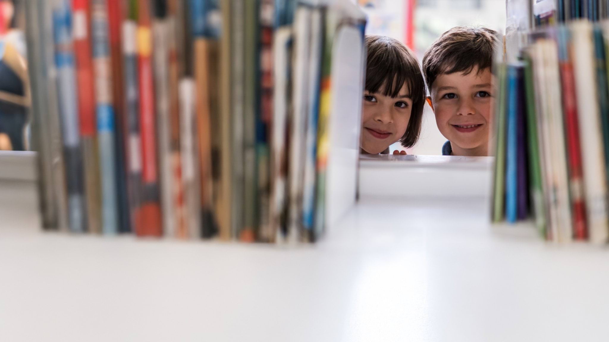 Education State banner Two children smiling and peering through a bookcase.