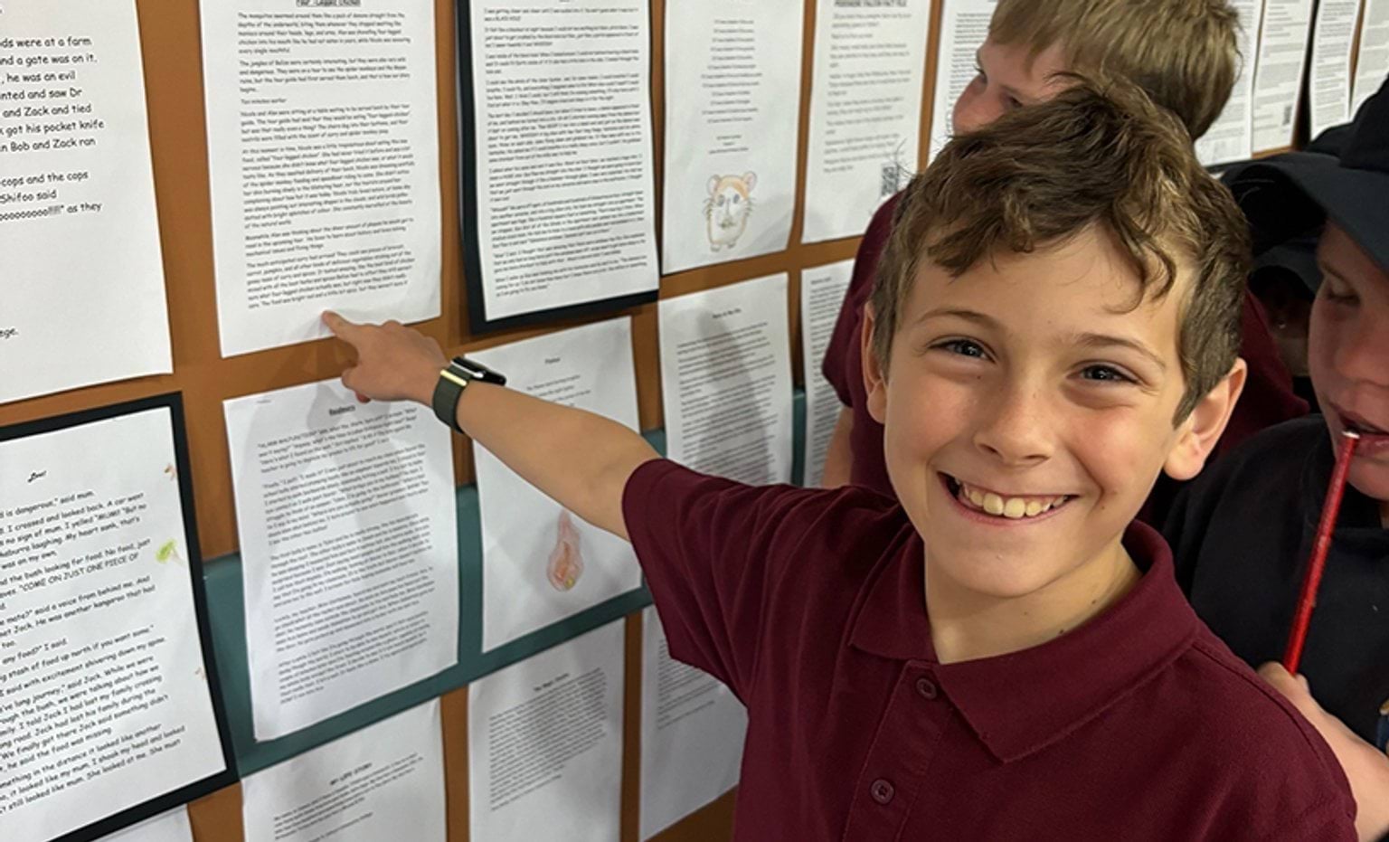 Budding authors showcase their skills A smiling boy in a burgundy shirt points at a wall displaying various printed pages.