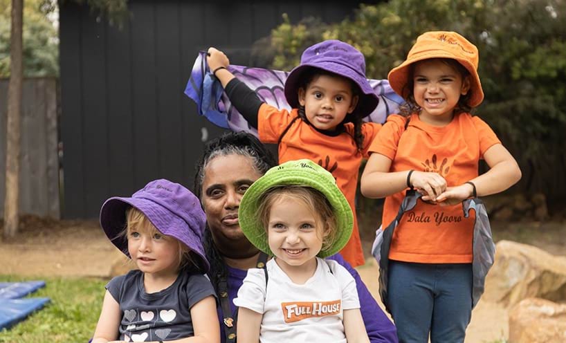 An early childhood professional and 5 children huddled together, smiling.