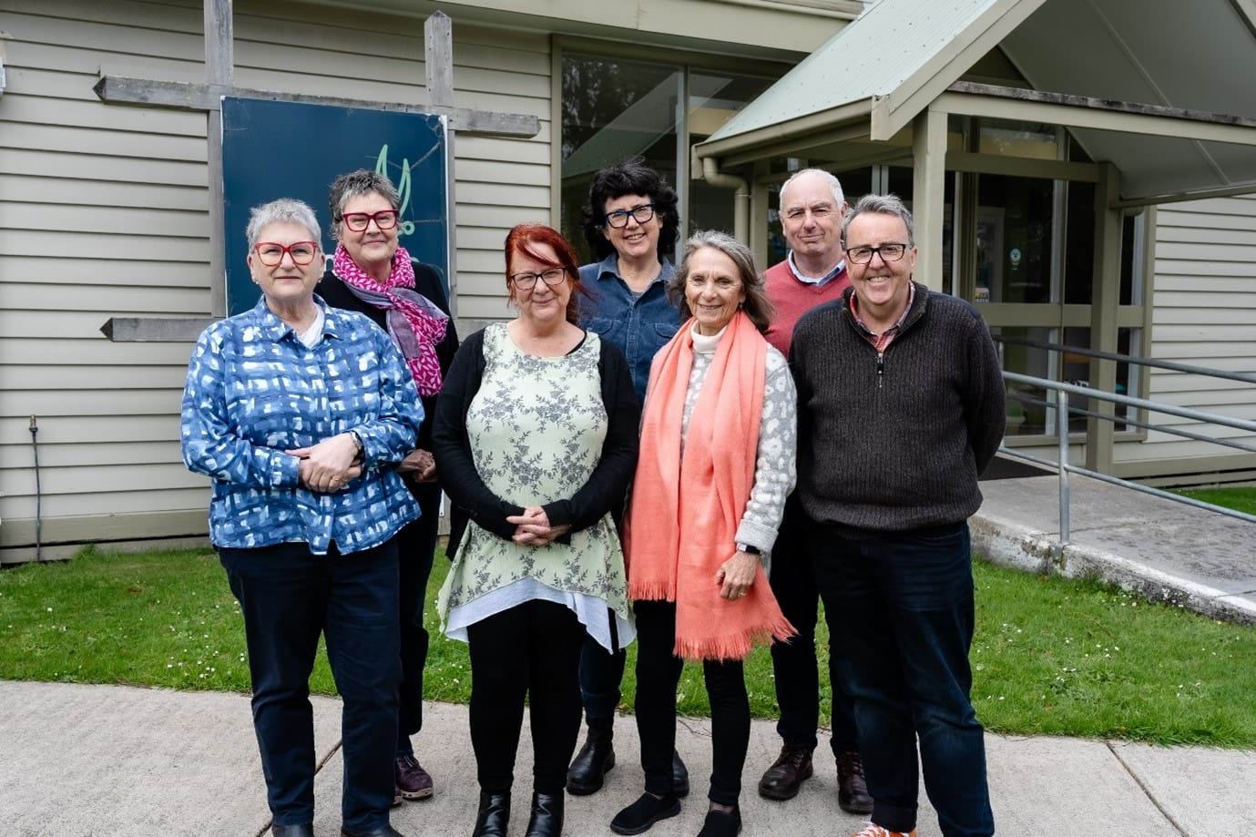 7 people standing, facing the camera and smiling, with a building in the background