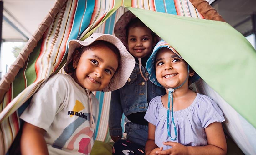 Three children wearing hats and smiling in a makeshift tent. 