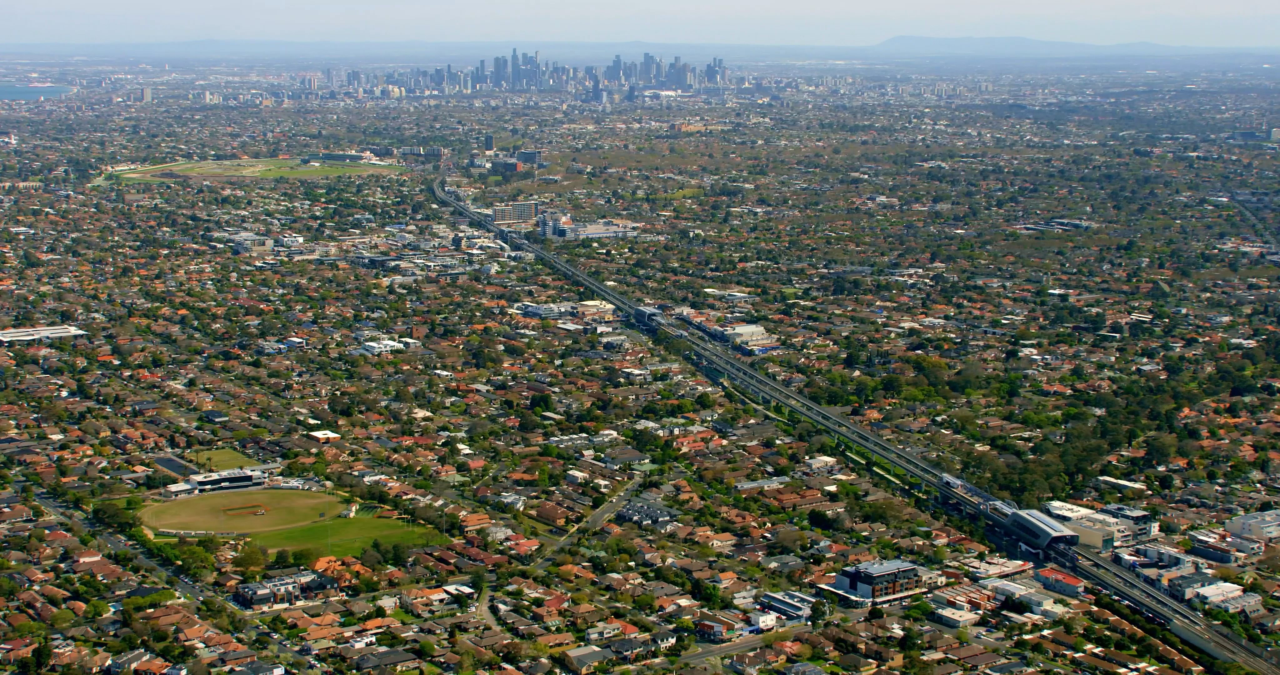 An aerial shot of the Melbourne sprawl, with low-rise suburbs in the foreground, and the CBD and mountain ranges in the background