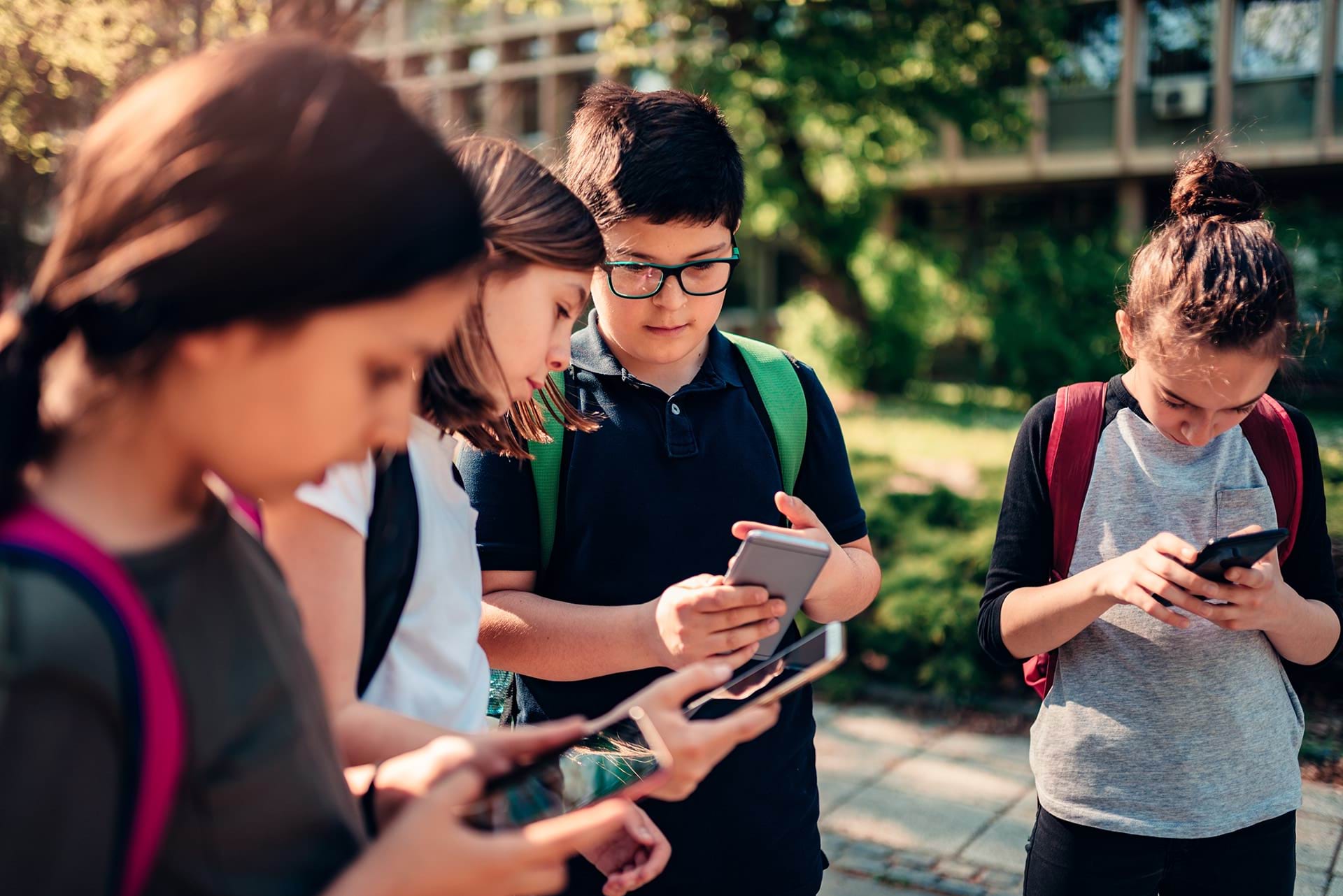 4 children 10-12 years old wearing backpacks look at their phones outside a school campus.