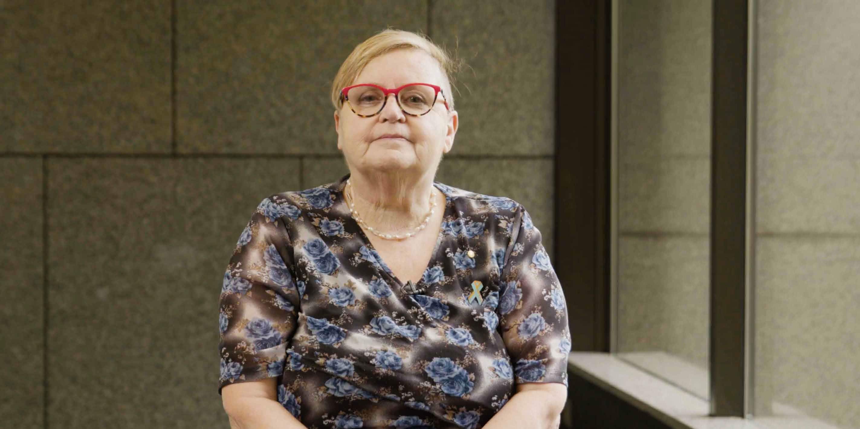 Tricia Malowney seated indoors, wearing red glasses and a floral blouse, looking directly at the camera.