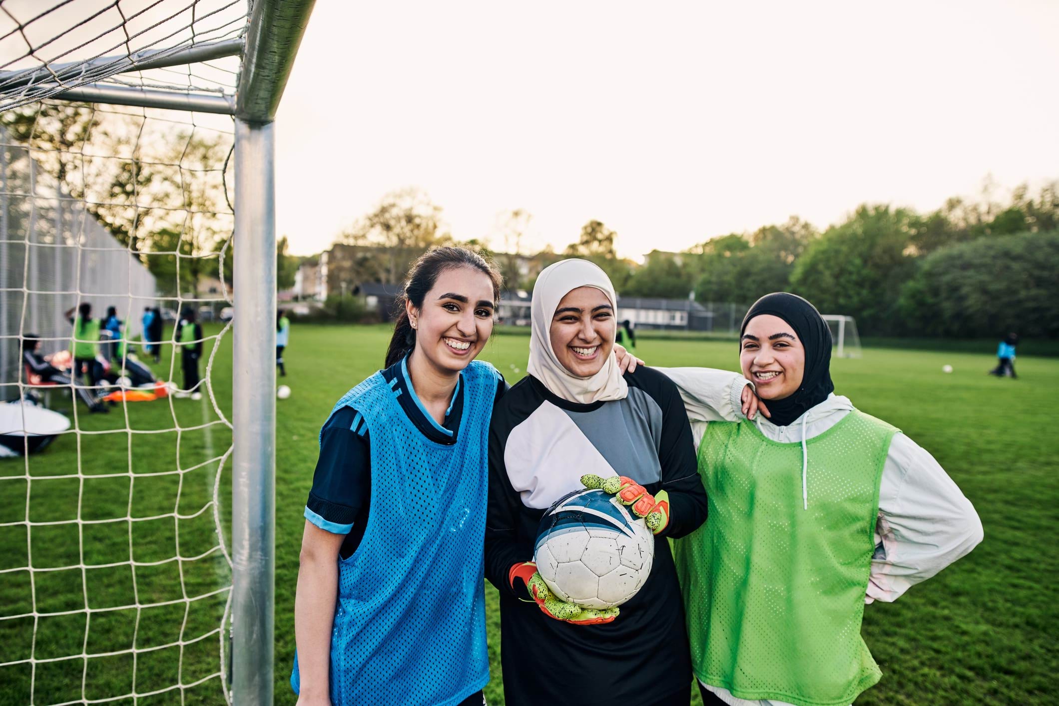 A group of three smiling girls in soccer gear stand next to a goal net. Two of the girls wear Islamic headscarves.