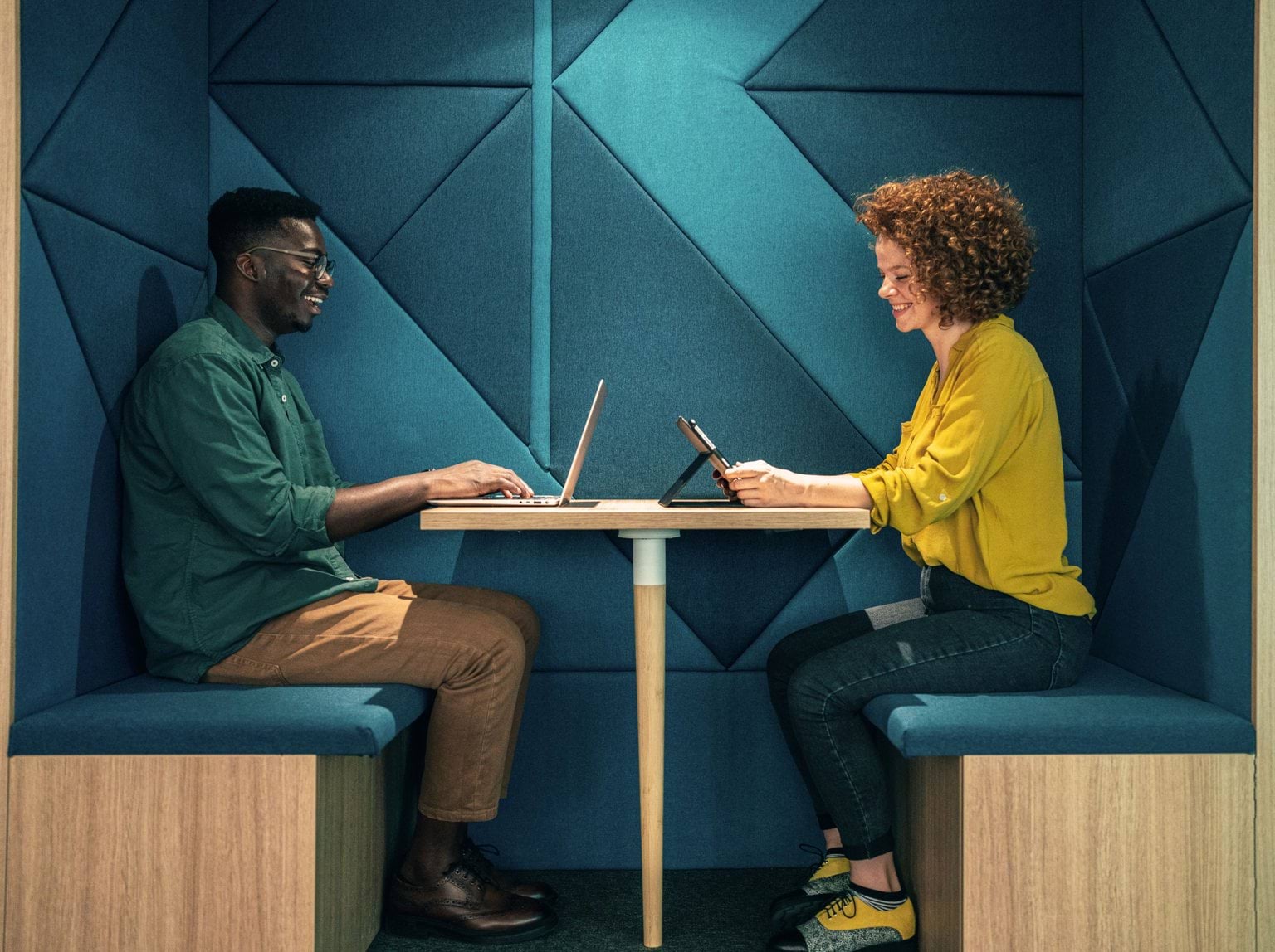 Two people sitting in a green booth with laptops.