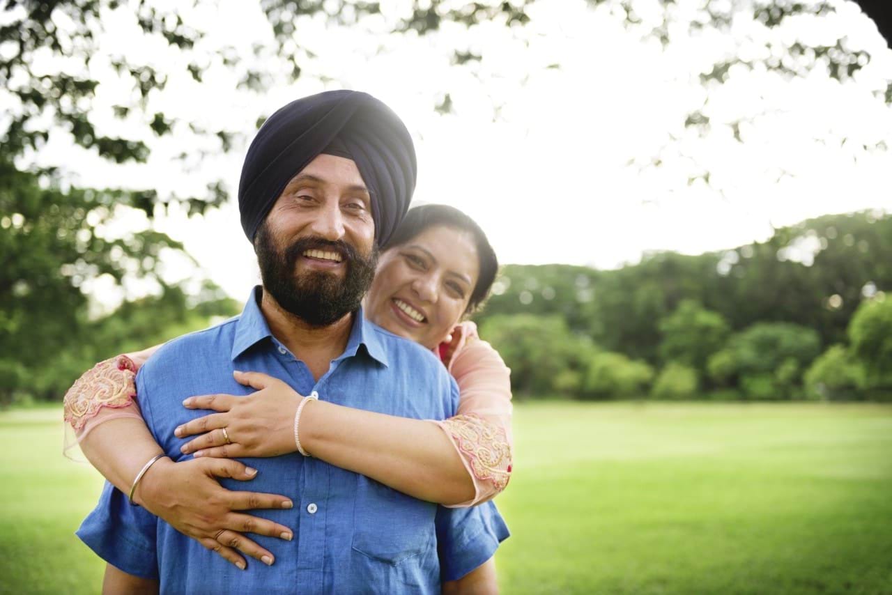 A smiling woman hugs from behind a smiling man who wears a black turban.