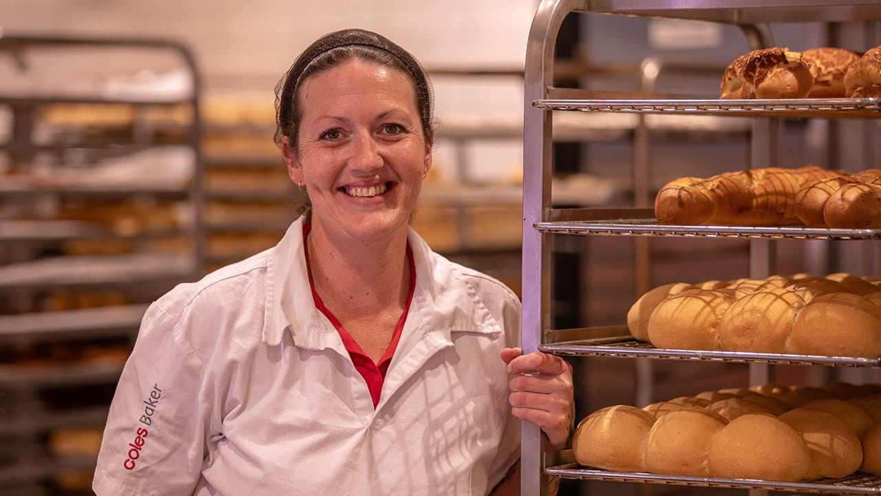 Student Satisfaction Survey participant Sarah standing in baker's uniform next to tray of bread rolls in kitchen work environment