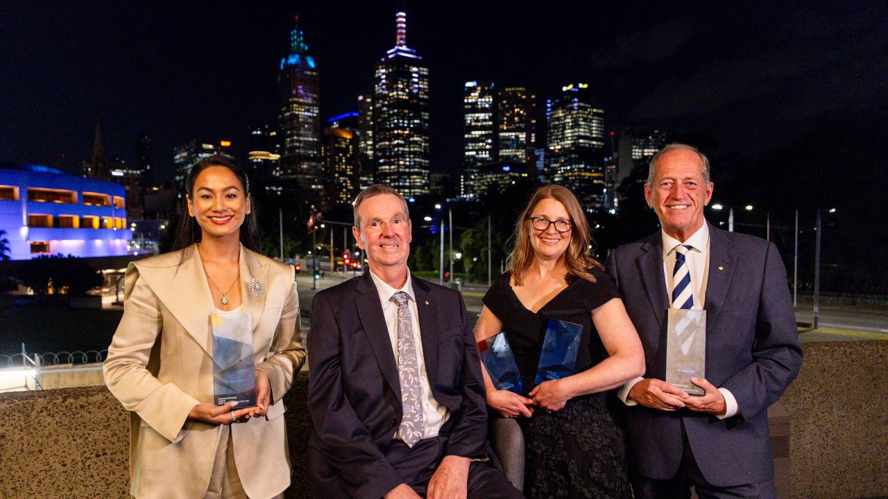 Four people smile in front of the camera holding awards, with the Melbourne skyline at night in the background