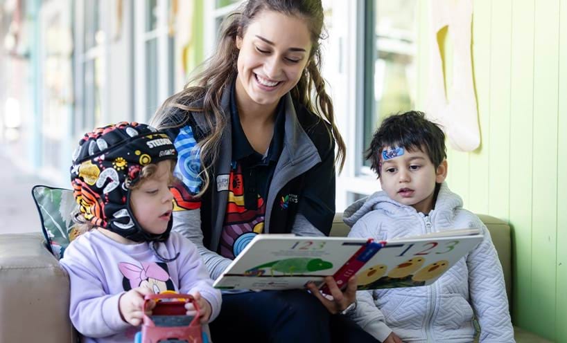 A smiling woman reads a book to two young children.