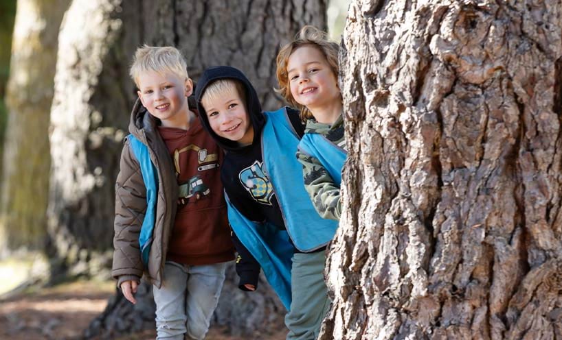 Three children in blue vests stand beside a tree, smiling and enjoying their time outdoors.