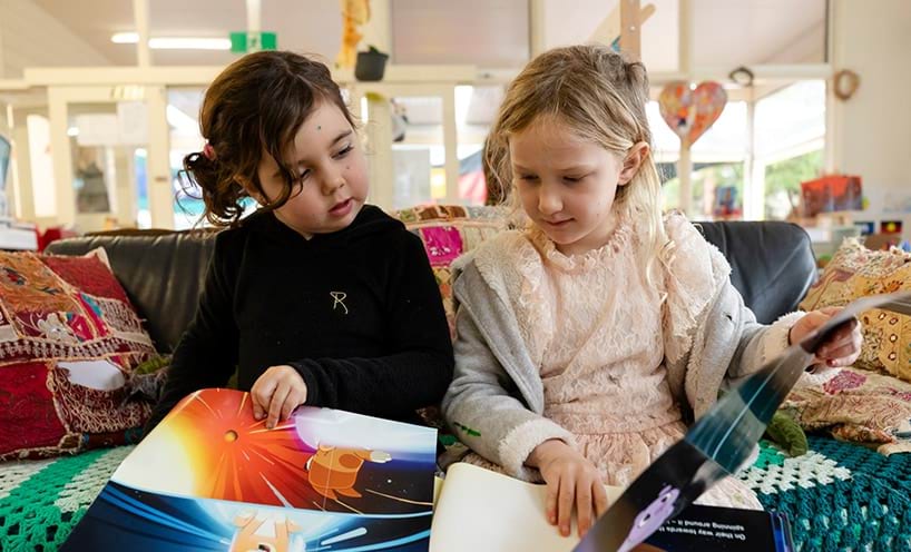 Two young girls sit on a couch reading their books together.