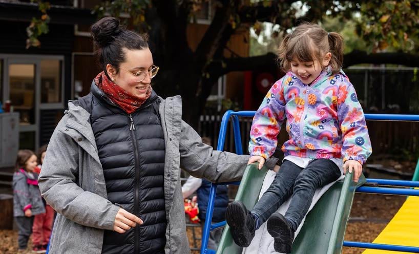 A woman standing next to a young girl on an outdoor slide.