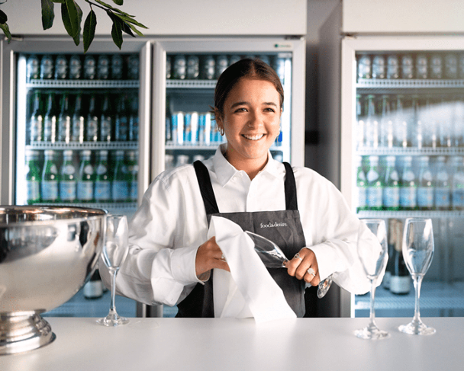 A person in a white shirt and apron smiles while drying wine glasses in a kitchen.