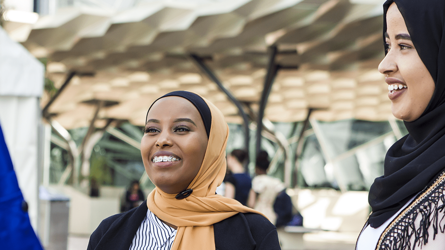 Two young women from African backgrounds wearing hijabs who are smiling and laughing in conversation with a third person, not featured in the image.
