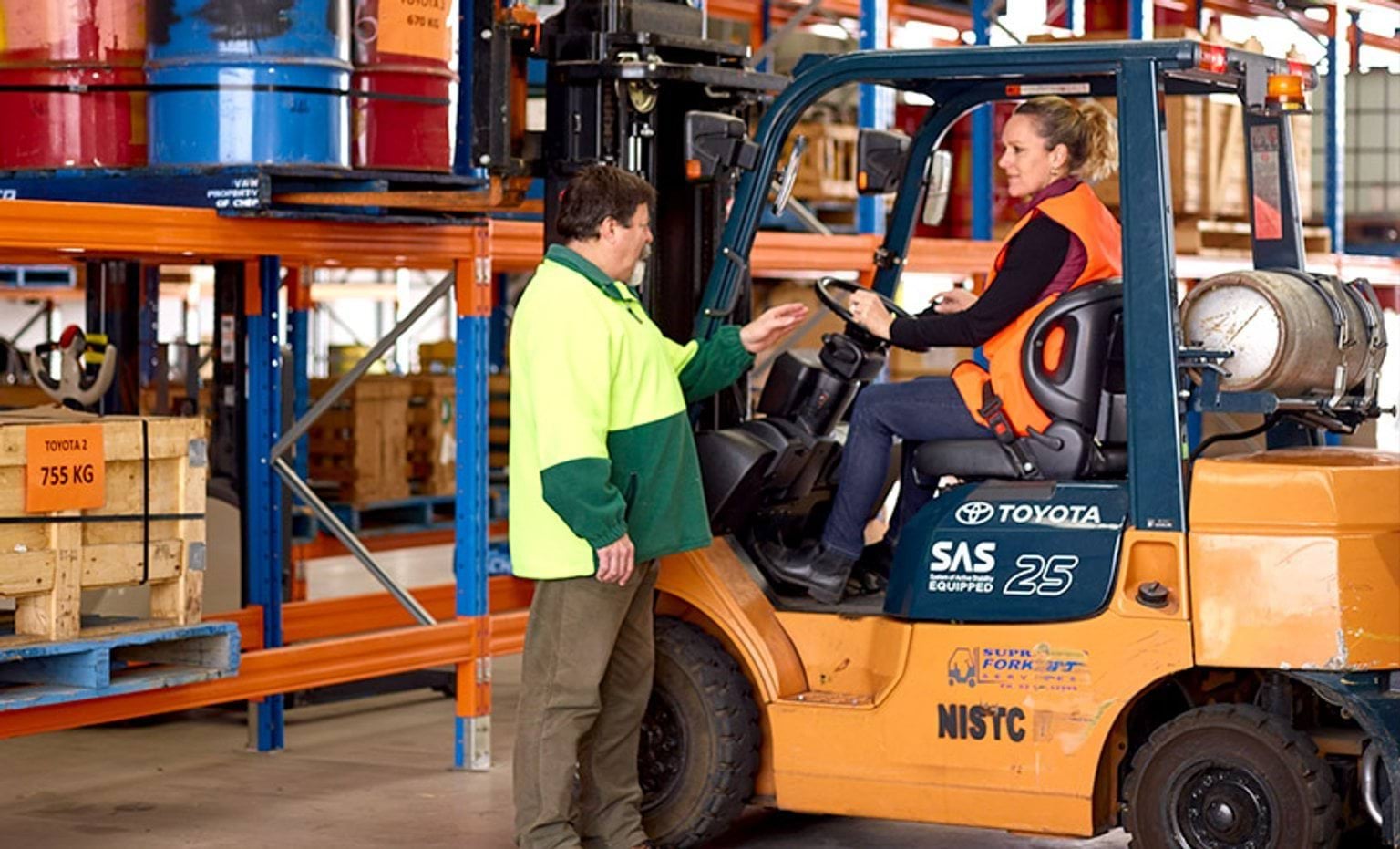 Two workers in a warehouse talking, one is sitting in a forklift