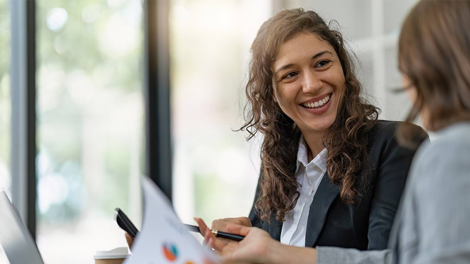 Two business people sitting at a desk talking and looking at a laptop and business docuemnts